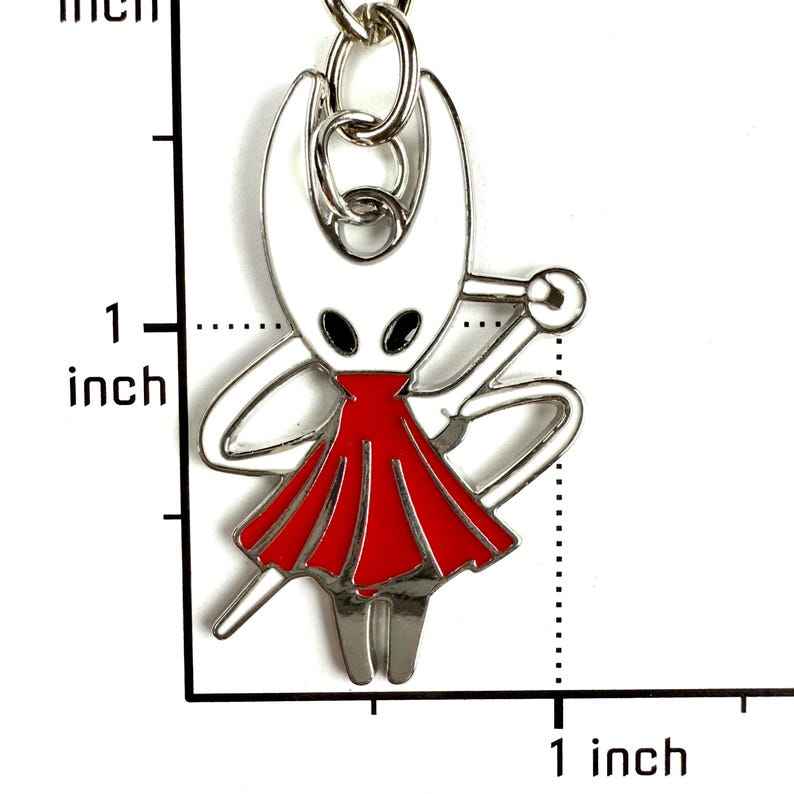 May include: A silver-toned metal keychain charm featuring a white character with black eyes and a red dress, holding a needle-like weapon. A ruler visible in the image indicates the charm measures approximately 1 inch in height and width.