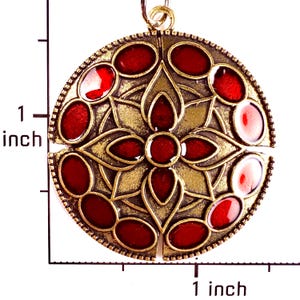 May include: A circular pendant with an antique brass finish featuring a central floral motif and twelve oval red enamel inlays. A ruler visible in the image indicates the pendant measures approximately 1 inch in diameter and 1 inch in height.