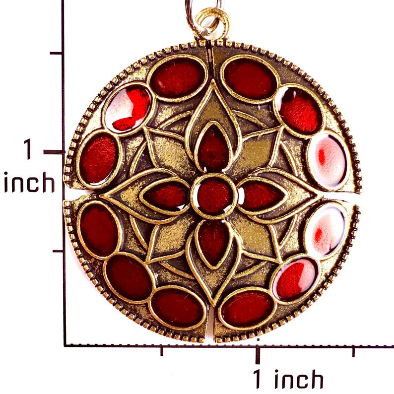 May include: A circular pendant with an antique brass finish featuring a central floral motif and twelve oval red enamel inlays. A ruler visible in the image indicates the pendant measures approximately 1 inch in diameter and 1 inch in height.