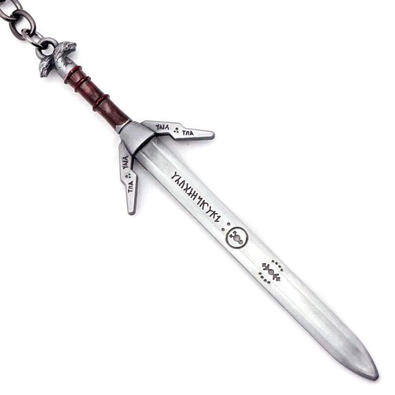 May include: A silver-toned metal sword keychain featuring a brown handle, a crossguard with engraved symbols, and a blade inscribed with runic characters. The pommel is shaped like a wolf head, and a metal chain is attached to the top of the hilt.