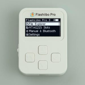 May include: A white rectangular electronic device labeled Flashiibo Pro with a digital screen displaying a menu including File Explorer, NTAG215 Slots, Manual & Bluetooth, and Settings. The device features four square navigation buttons below the screen.