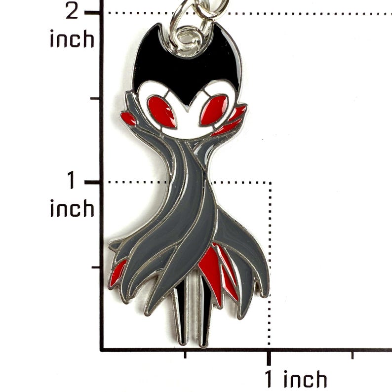 May include: A metal enamel keychain shaped like a stylized character with a black head, white face, and red eyes. The body is draped in dark gray fabric-like layers with red accents. A ruler shows the item is approximately 2 inches in total height.