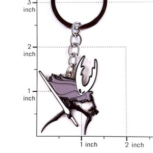 May include: A metal keychain featuring a character with a white mask and a light purple cloak, holding a thin white sword. The keychain is displayed against a grid with markings for 1, 2, and 3 inches, showing the character is about 1.5 inches tall.
