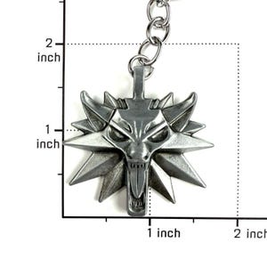 May include: A silver-toned metal wolf head keychain pendant featuring a stylized, angular design with a protruding tongue. The pendant is positioned against a measurement grid, showing a width and height of approximately 1.5 inches.