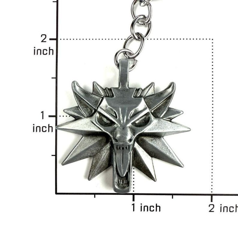 May include: A silver-toned metal wolf head keychain pendant featuring a stylized, angular design with a protruding tongue. The pendant is positioned against a measurement grid, showing a width and height of approximately 1.5 inches.
