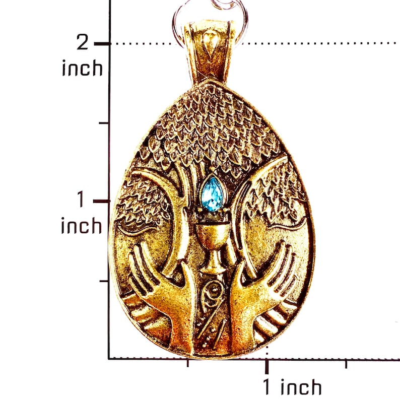May include: A gold-toned oval pendant featuring a raised relief design of two hands cradling a chalice beneath a tree, accented with a single blue teardrop-shaped stone. The pendant measures approximately 2 inches in height and 1 inch in width.