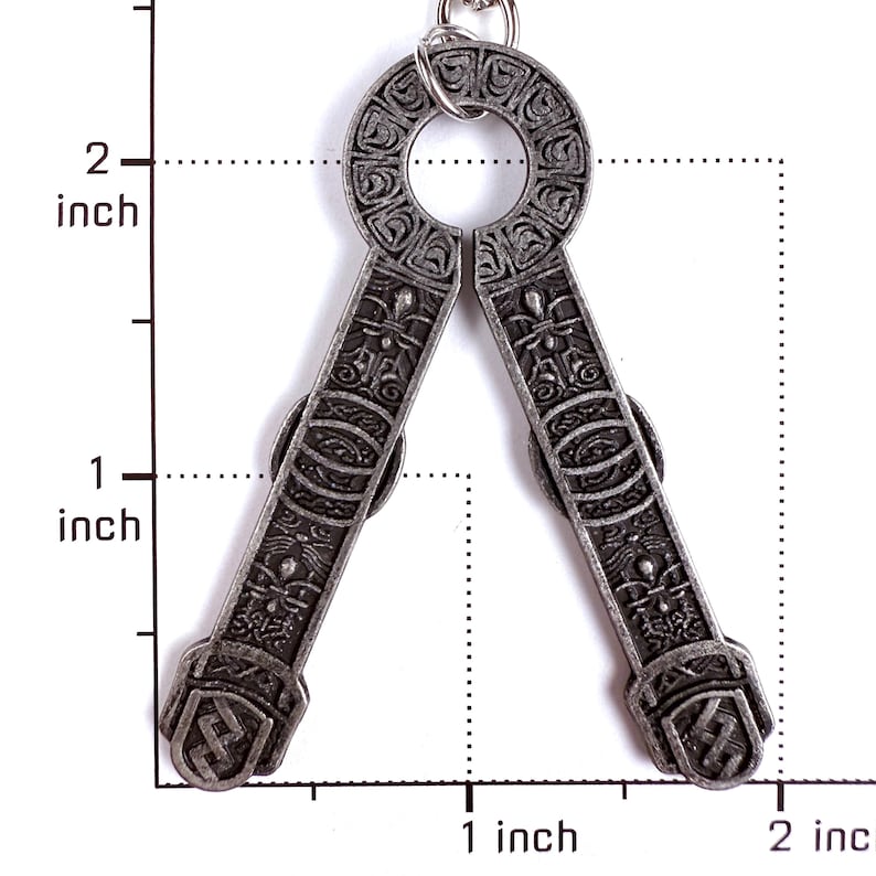 May include: A silver-toned metal pendant featuring an intricate embossed pattern and a circular top loop. The pendant is positioned against a scale showing a height of approximately 2 inches and a width of approximately 1.5 inches.
