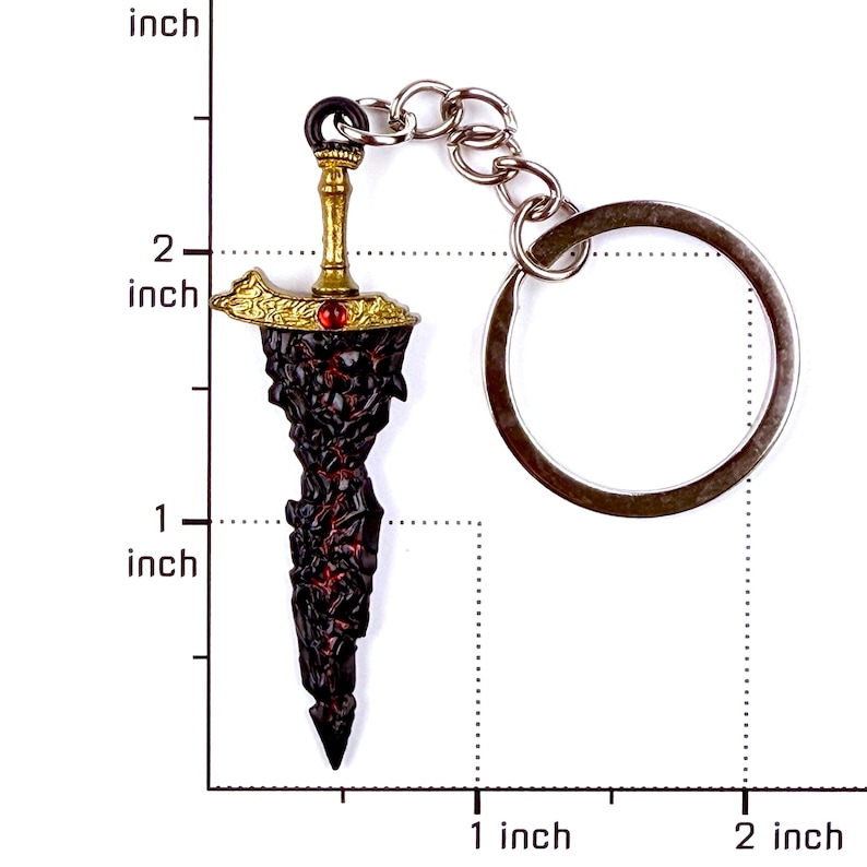 May include: A sword-shaped keychain featuring a gold-colored hilt with a small red gem and a textured, dark, jagged blade. The keychain is positioned against a ruler showing the sword length is approximately 1.5 inches from tip to hilt.