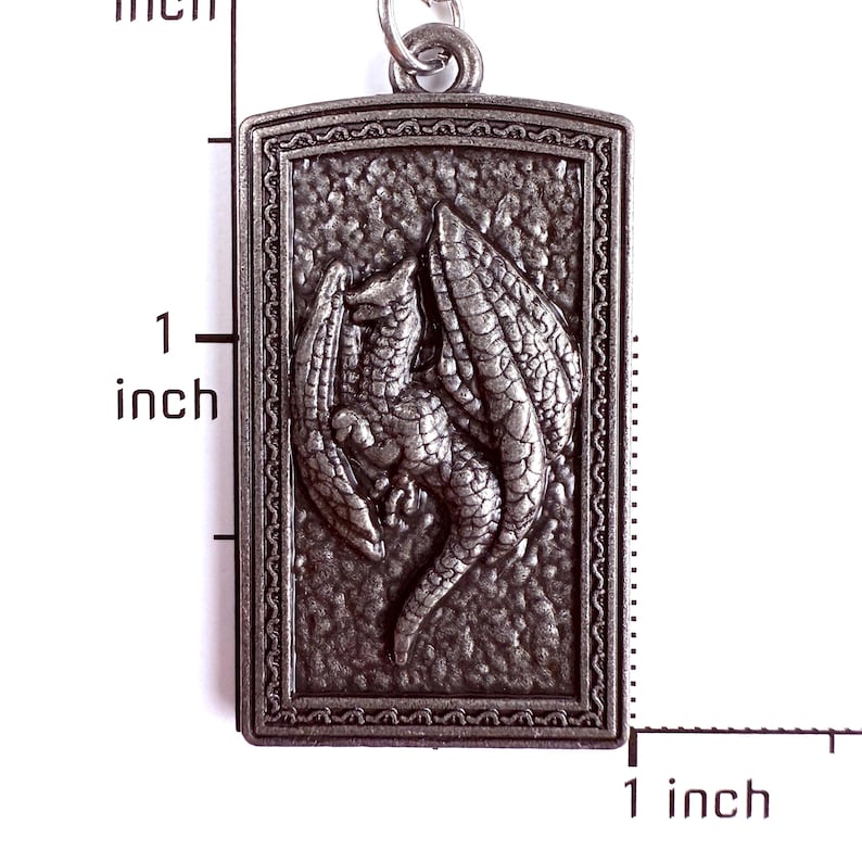 May include: A rectangular silver-toned metal pendant featuring a raised relief of a dragon with wings spread. The pendant is positioned next to a ruler showing a height of approximately 1.5 inches and a width of approximately 0.75 inches.