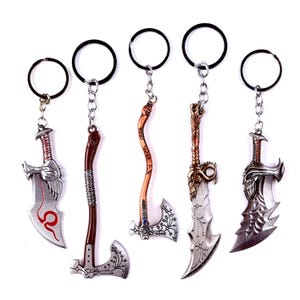 May include: Five metal keychains featuring miniature weapon designs. The keychains include swords and axes with intricate details, attached to black key rings. The weapons have silver, brown, and bronze tones, with some featuring red accents.