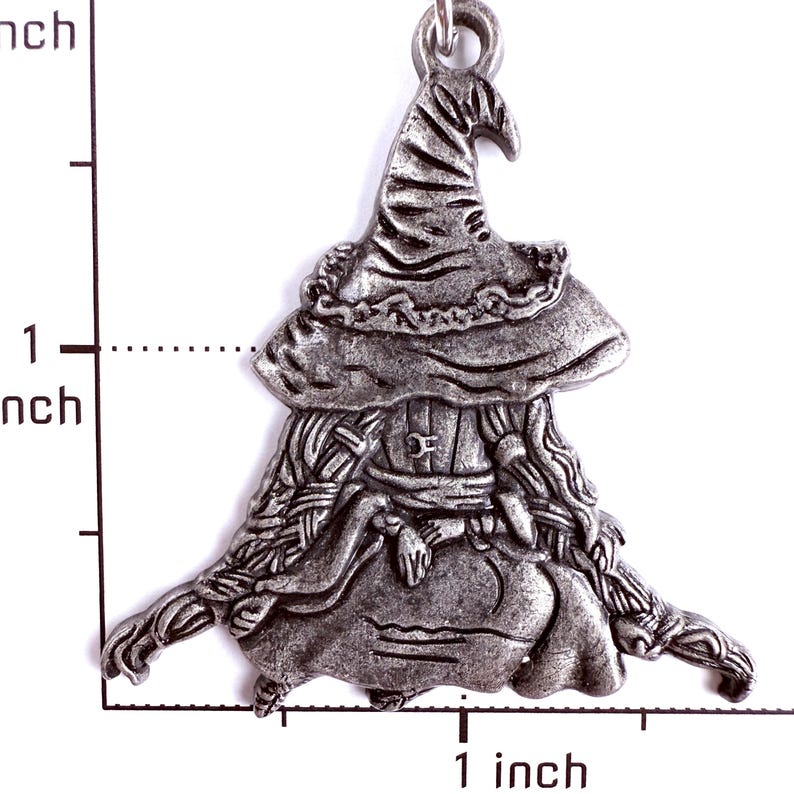 May include: A silver-toned metal charm shaped like a figure wearing a wide-brimmed pointed hat and a long garment. The charm is positioned against a white background with a ruler showing a height and width of approximately 1.5 inches.