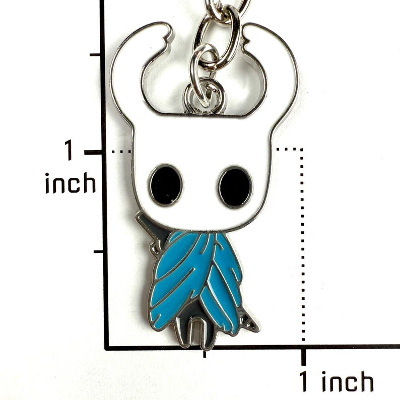 May include: A metal keychain charm featuring a white character with large black oval eyes and a bright blue cloak. The charm is attached to a silver metal ring. A black graphic scale indicates the charm is approximately 1 inch in height.