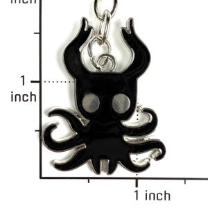 May include: A black enamel keychain charm shaped like a stylized creature with two large horns and multiple tentacles, featuring two circular silver eyes. A ruler overlay indicates the charm measures approximately 1 inch in height and width.