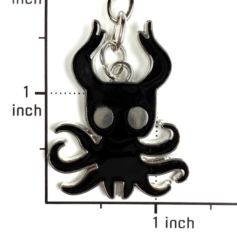 May include: A black enamel keychain charm shaped like a stylized creature with two large horns and multiple tentacles, featuring two circular silver eyes. A ruler overlay indicates the charm measures approximately 1 inch in height and width.