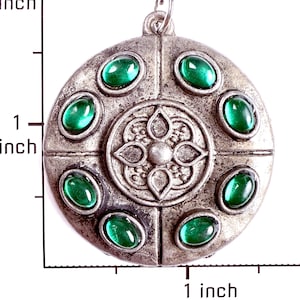 May include: A round, silver-toned metal pendant featuring a central embossed floral motif surrounded by six oval, cabochon-cut green stones. A ruler visible on the left and bottom indicates the pendant measures approximately 1.5 inches in diameter.