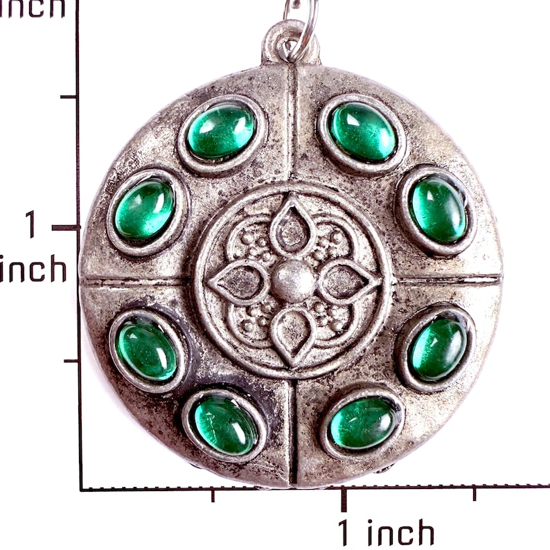 May include: A round, silver-toned metal pendant featuring a central embossed floral motif surrounded by six oval, cabochon-cut green stones. A ruler visible on the left and bottom indicates the pendant measures approximately 1.5 inches in diameter.