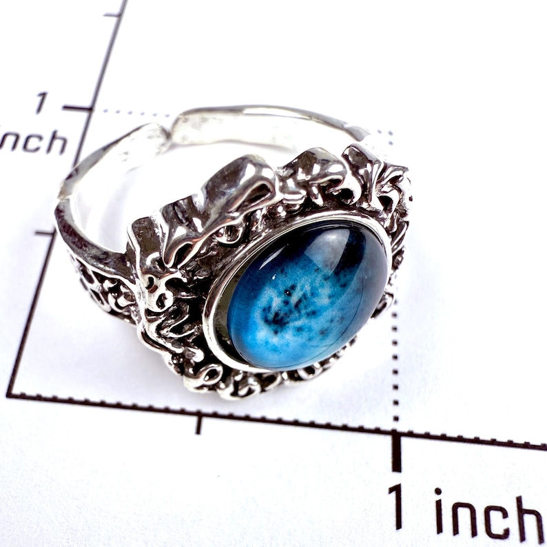 May include: A silver-toned ring featuring an oval, deep blue cabochon with a mottled, cloud-like pattern. The metal band has an intricate, textured, and raised relief design. A ruler visible in the image shows the ring is approximately 1 inch wide.