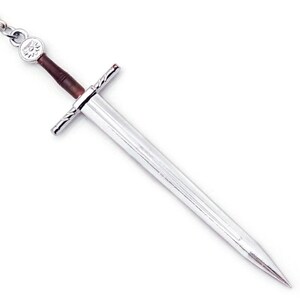 May include: A silver-toned metal sword keychain featuring a long, pointed blade, a crossguard with textured detailing, and a dark brown cylindrical handle. At the top of the handle, a circular silver emblem displays a wolf head design.