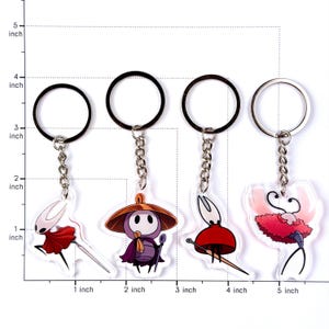 May include: Four acrylic keychains featuring characters from Hollow Knight, each attached to a silver metal ring and chain. The characters include Hornet, a purple figure with a wide hat, a small red-cloaked figure, and a pink creature. A ruler grid shows sizes in inches.