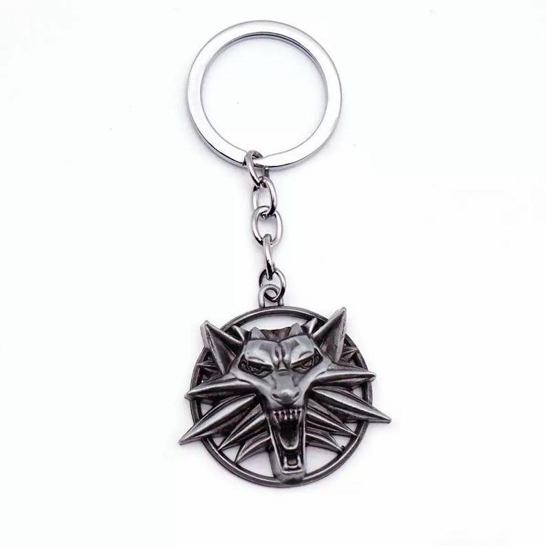 May include: A silver-toned metal keychain featuring a circular pendant with a detailed, stylized wolf head design. The wolf head is centered within a thin circular frame, and the pendant is attached to a standard split ring by a short chain.