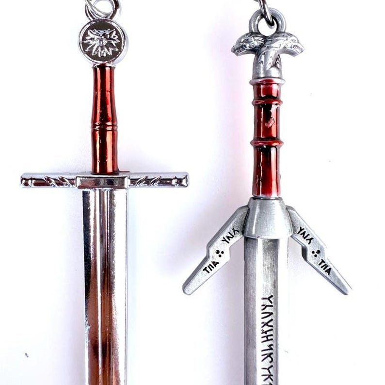 May include: Two metal sword pendants featuring silver-toned blades and deep red handles. The left pendant has a circular wolf emblem at the pommel. The right pendant features a dual-wolf head pommel and engraved runic text along the blade and crossguard.