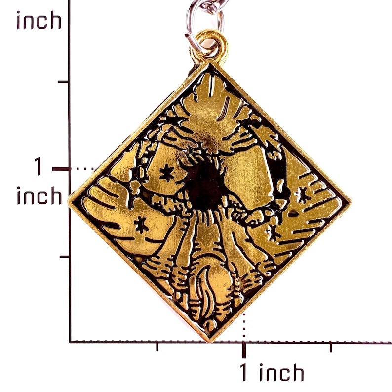 May include: A diamond-shaped gold-toned metal pendant featuring a raised relief design of a central figure surrounded by radiating lines and small star shapes. A ruler visible on the left and bottom indicates the pendant is approximately 1 inch tall.