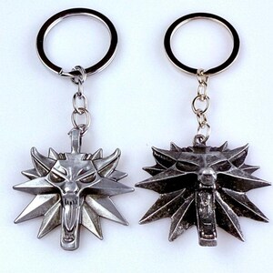 May include: Two silver-toned metal keychains featuring a stylized wolf head emblem with sharp, radiating points. Each pendant is attached to a circular split ring by a short metal chain. The wolf designs display detailed facial features with an open mouth.