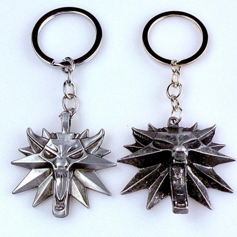 May include: Two silver-toned metal keychains featuring a stylized wolf head emblem with sharp, radiating points. Each pendant is attached to a circular split ring by a short metal chain. The wolf designs display detailed facial features with an open mouth.