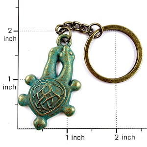 May include: A bronze-toned metal keychain featuring a turtle-shaped charm with a weathered teal and gold patina finish. The charm is attached to a circular split ring by a short chain. A ruler overlay indicates the charm is approximately 2 inches tall.