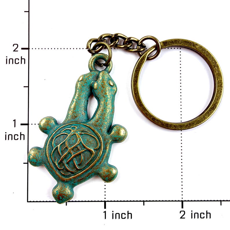 May include: A bronze-toned metal keychain featuring a turtle-shaped charm with a weathered teal and gold patina finish. The charm is attached to a circular split ring by a short chain. A ruler overlay indicates the charm is approximately 2 inches tall.