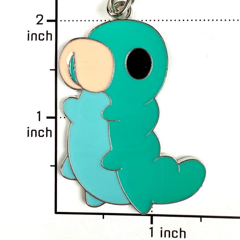 May include: A teal and light blue enamel charm shaped like a cartoon caterpillar with a peach-colored head and a black eye. The charm is positioned against a ruler showing a height of approximately 1.75 inches and a width of 1 inch.