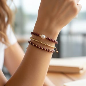 May include: Three bracelets stacked on a wrist. The top bracelet has dark red beads and a large white pearl. The middle bracelet is gold, and the bottom bracelet has dark red beads. The bracelets are on a wrist with a white shirt in the background.