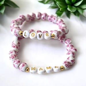 May include: Two white beaded bracelets with pink floral patterns. The top bracelet spells out "LOVE" in gold letters and the bottom bracelet spells out "MAMA" in gold letters.
