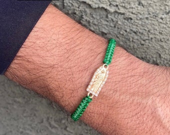 Puede incluir: Pulsera de cuerda verde con un dije de oro que representa una figura religiosa. El dije está adornado con diamantes de imitación transparentes. La pulsera está atada con un nudo.