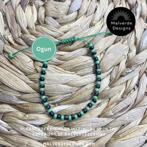 May include: A green and black beaded bracelet with the word "Ogun" in a green circle. The bracelet has a sliding knot closure. The text "Malverde Designs" is in a black circle. The text "MALVERDEDESIGNS.COM" is at the bottom.