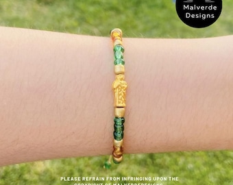 Puede incluir: Una pulsera de cuentas doradas y verdes con una pequeña estatua dorada en el centro. La pulsera está en el brazo de una persona. La imagen incluye el texto "Malverde Designs" y "PLEASE REFRAIN FROM INFRINGING UPON THE COPYRIGHT OF MALVERDEDESIGNS."