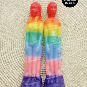 Op de afbeelding: Twee regenboogkleurige figuurkaarsen. Elke kaars is een menselijke vorm met een rode kop, die overgaat in oranje, geel, groen, blauw, roze en paars. De kaarsen hebben de tekst "MALVERDEDESIGNS" erop. De achtergrond is een beige geweven oppervlak.