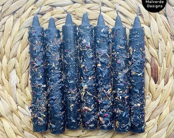 May include: A collection of seven black spell candles, decorated with dried herbs in various colours, are displayed on a woven mat. The candles have a textured finish and wicks. The image includes the text "Malverde Designs" and a copyright notice.