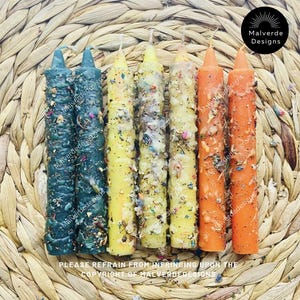 Puede incluir: Seis velas de hechizo coloridas en tonos verdes, amarillos y naranjas, adornadas con hierbas secas y botánicos. Las velas están dispuestas sobre una superficie tejida y texturizada. La imagen incluye el texto "Malverde Designs" y un aviso de derechos de autor.