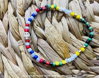 May include: A colorful beaded bracelet with a white skull charm. The beads are red, white, blue, yellow, green, and black. The bracelet is made by Malverde Designs.