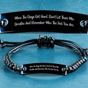 May include: Black bracelet with a rectangular metal plate engraved with the text: "When The Days Get Hard. Don't Let Them Win. Breathe And Remember Who The F*ck You Are." The bracelet is made of black woven cord.