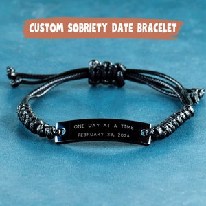 May include: Black bracelet with a black metal tag that reads "One Day At A Time" and "February 28, 2024".