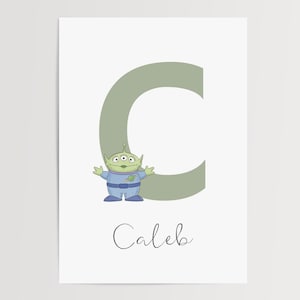 May include: A print featuring a green alien character from Toy Story, beside a large green letter "C". The name "Caleb" is written in cursive below the letter and character. The background is white.