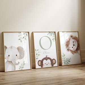 May include: Three framed nursery art prints featuring watercolor illustrations of animals. The prints include an elephant, a monkey with the name "Olive", and a lion. Each print has a light wood frame and botanical accents.