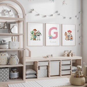 May include: Three framed prints featuring Toy Story characters and a pink letter "G" with the name "Grace". The prints are displayed above a light wood storage unit with white storage bins. The room is decorated with a string of lights and decorative items.