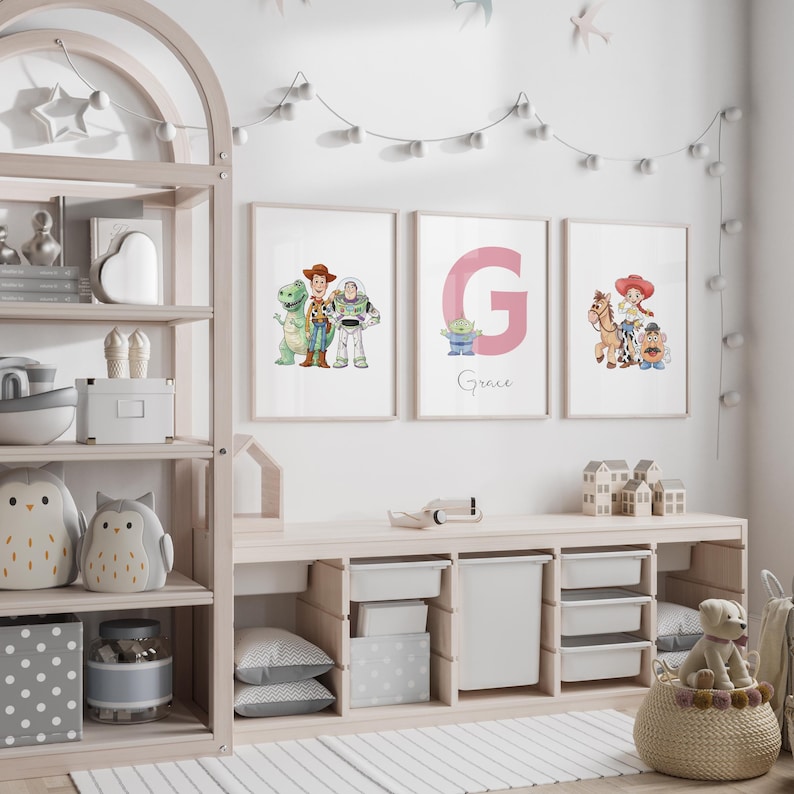 May include: Three framed prints featuring Toy Story characters and a pink letter "G" with the name "Grace". The prints are displayed above a light wood storage unit with white storage bins. The room is decorated with a string of lights and decorative items.
