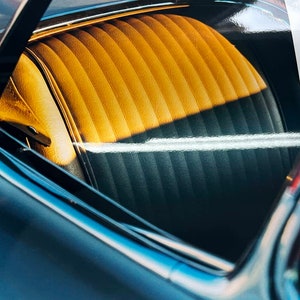 May include: A close-up of the interior of a classic car. The back seat is visible through the rear window. The seat is upholstered in a yellow and black striped fabric. The car's interior is sleek and stylish.