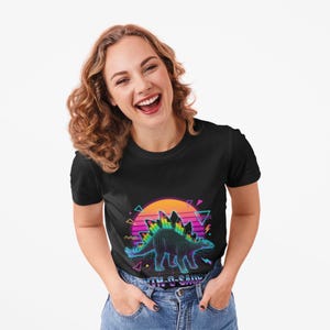 May include: Black t-shirt featuring a neon-outlined Stegosaurus design with a sunset backdrop and geometric shapes. The text "Ninth-O-Saur" is below the dinosaur. The design has a retro, 80s-inspired aesthetic.