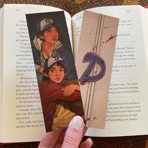 May include: Two illustrated bookmarks featuring a character wearing a baseball cap. One bookmark has a large letter "D" design. The bookmarks are placed in an open book with red edges.