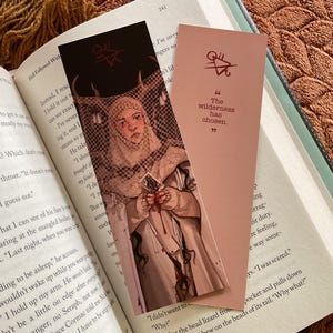 Op de afbeelding: Twee bladwijzers liggen op een open boek. Eén toont een illustratie van een persoon in een wit gewaad, die een kaart vasthoudt. De andere is roze met de tekst "The wilderness has chosen."